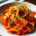 Close-up of a vibrant raw carrot salad made with shaved carrot ribbons, topped with sesame seeds and chopped green onions.