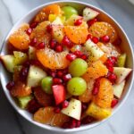 Close-up of a bright winter fruit salad featuring mandarin segments, green grapes, apple chunks, and pomegranate seeds.