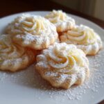 A close-up of several delicious whipped shortbread cookies, piped into rosettes and dusted heavily with powdered sugar.